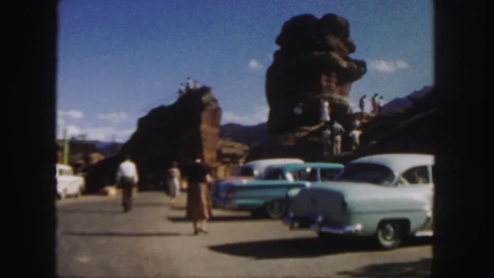 1958 Colorado visitor area with people, parked mid-century cars, and large rock formations under blue sky.
