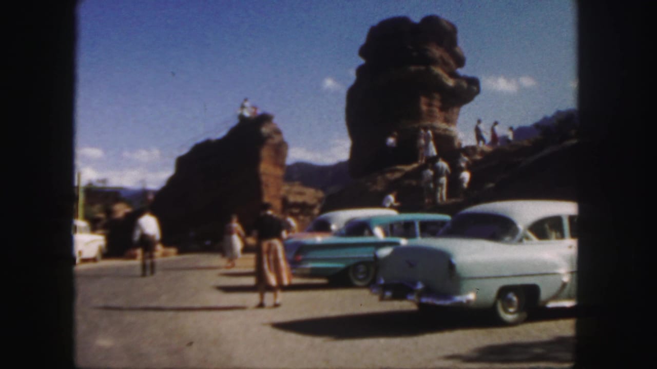 Tourists and classic cars beside large red rock formations under a blue sky.
