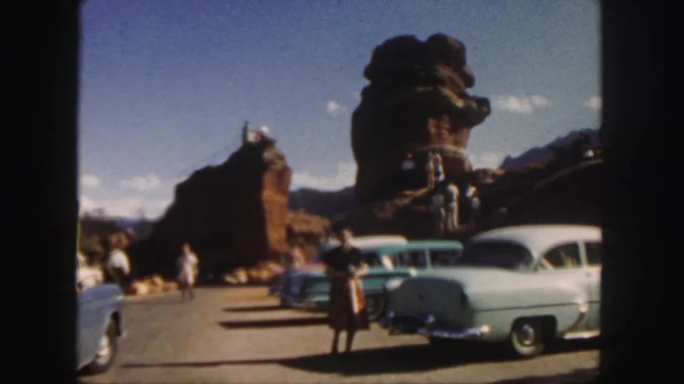 Colorado home-movie frame from 1958 with a woman in period dress near parked cars and red rock scenery.