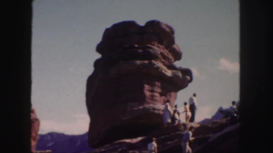 1958 Colorado 8mm frame showing visitors on and below a massive red sandstone formation with distant mountains behind.