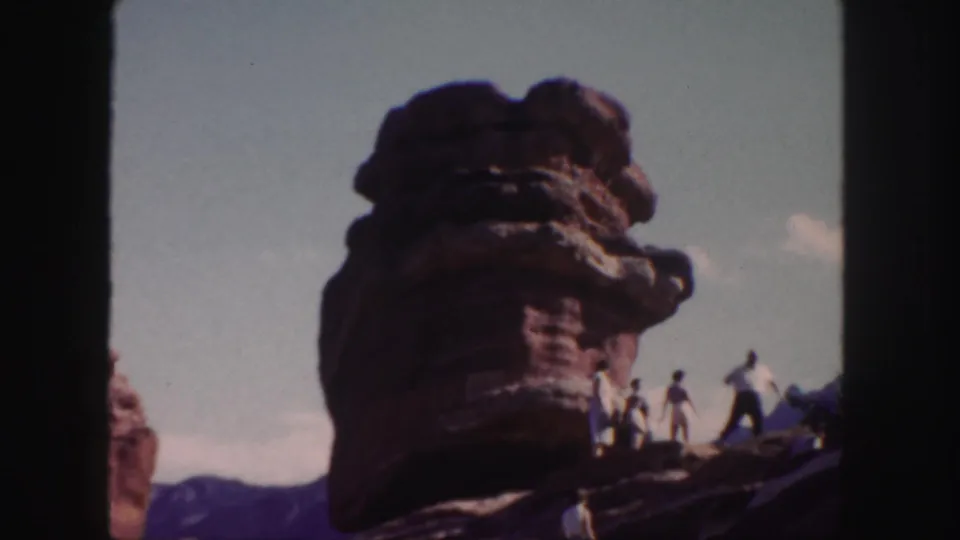 Visitors in 1958 Colorado home-movie footage walk and stand near a sculpted red rock formation beneath open sky.