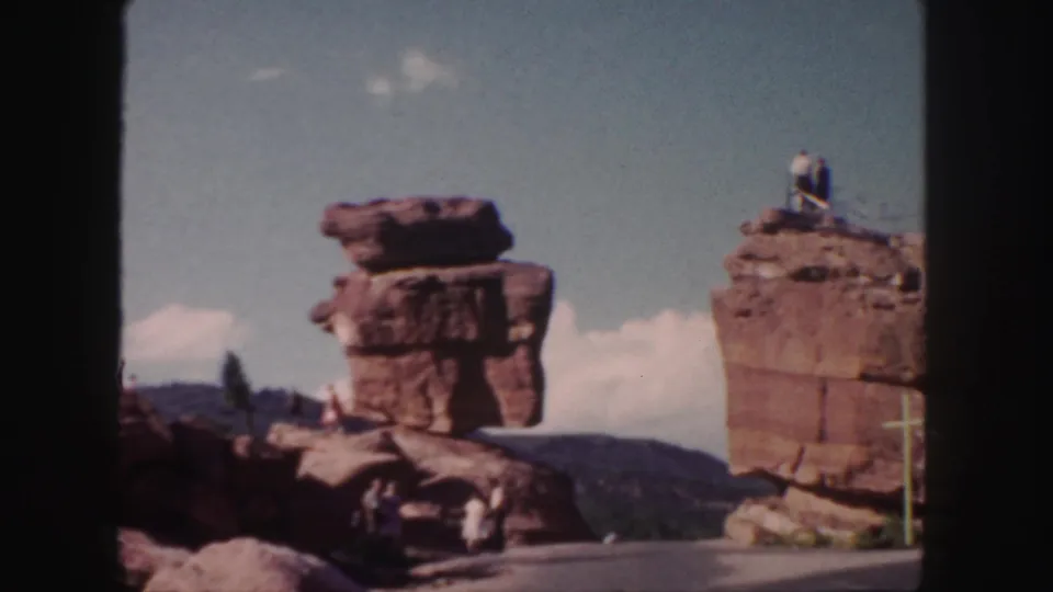 1958 Colorado home-movie frame with balanced red rocks, a paved overlook area, distant hills, and a man atop the right formation.