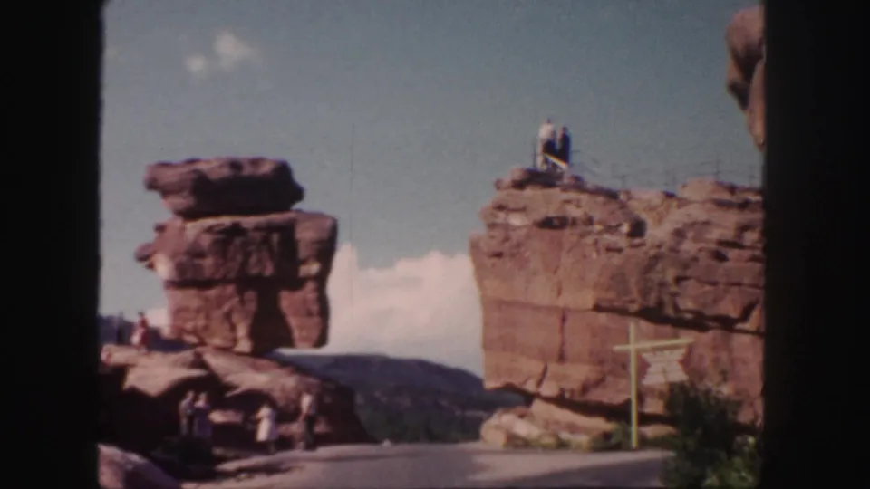 1958 Colorado 8mm frame showing tourists around red rock formations, a paved path, and a person standing on the elevated right ledge.