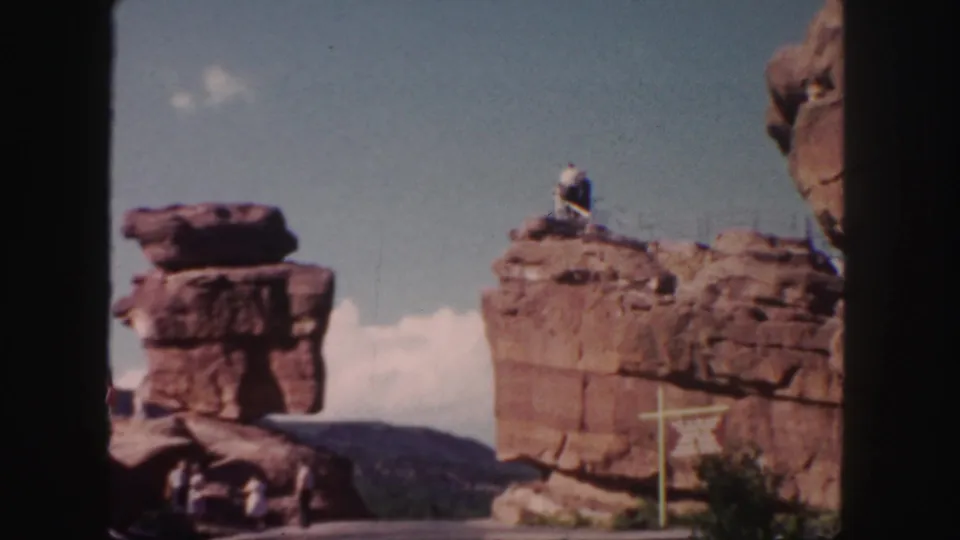 1958 Colorado home-movie frame of red rock formations, visitors at the base, a person atop the right ledge, and mountains beyond.