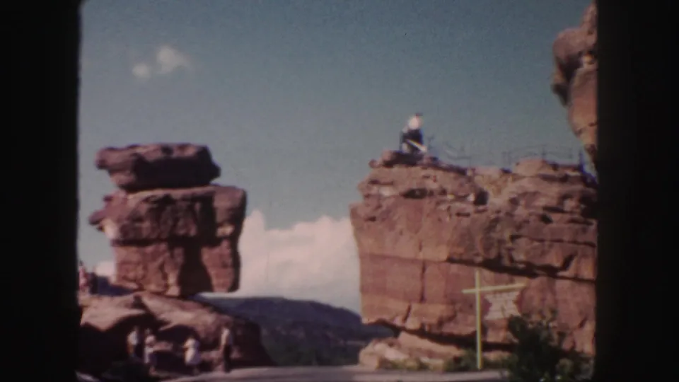 1958 Colorado 8mm frame showing red rock formations, a white roadside signpost, shrubs, and a small distant visitor near the rocks.