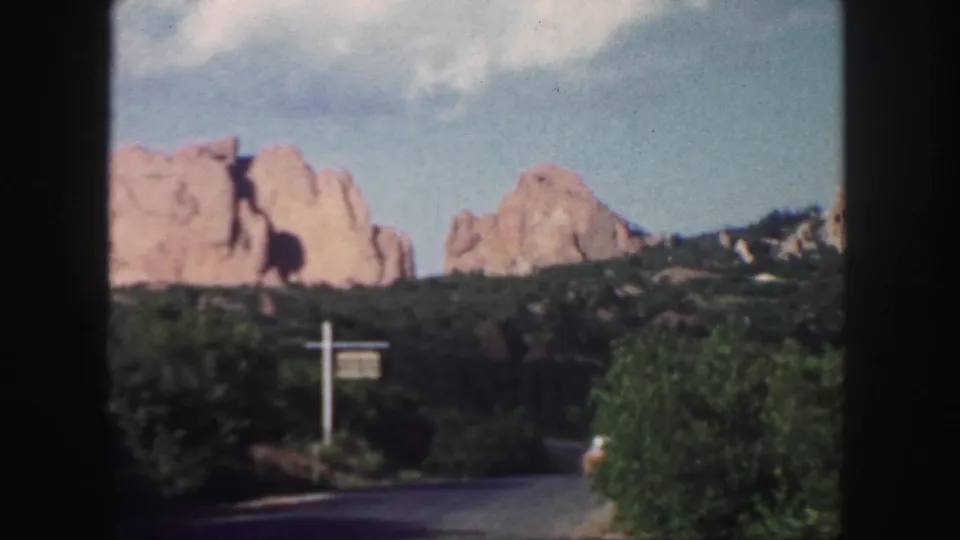 1958 Colorado 8mm frame showing a roadside signpost, paved road, dense greenery, and red rock formations across the hillside.