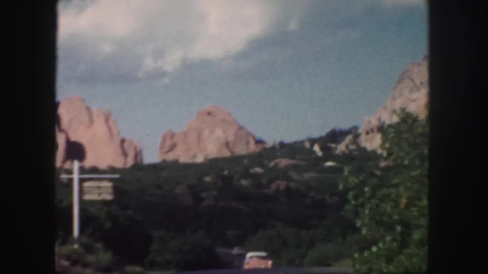 1958 Colorado frame with red rock formations, roadside signpost, green shrubs, a paved road, and a partial car roof at the lower edge.