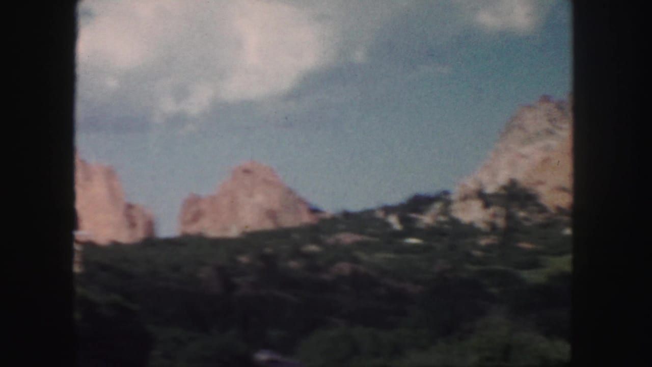 Tan rock cliffs above dark green hillside under a blue, clouded sky.