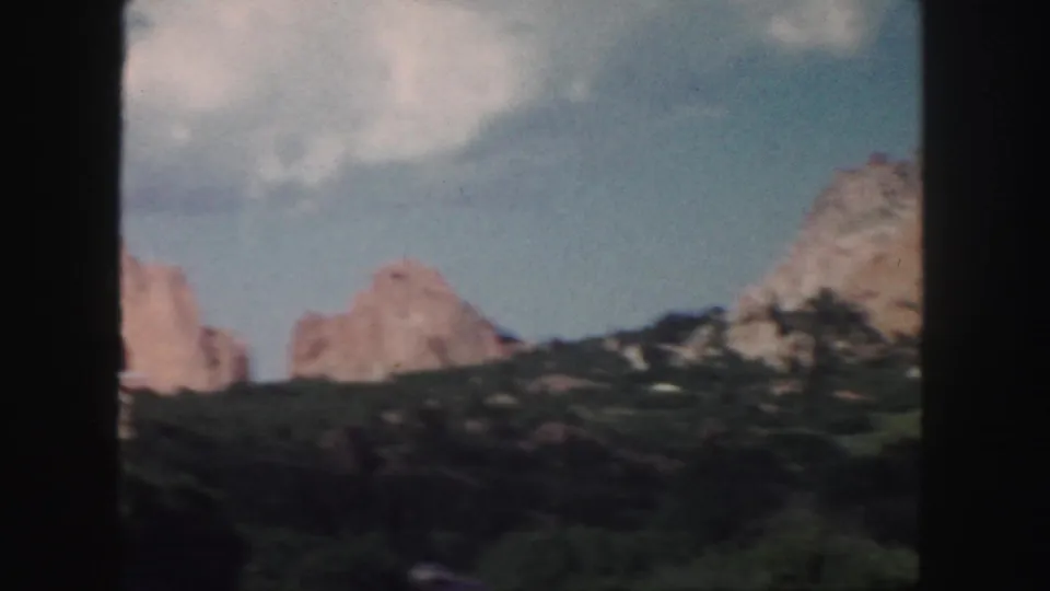 1958 Colorado 8mm frame showing tan mountain cliffs, green hillside, and blue sky with scattered clouds.