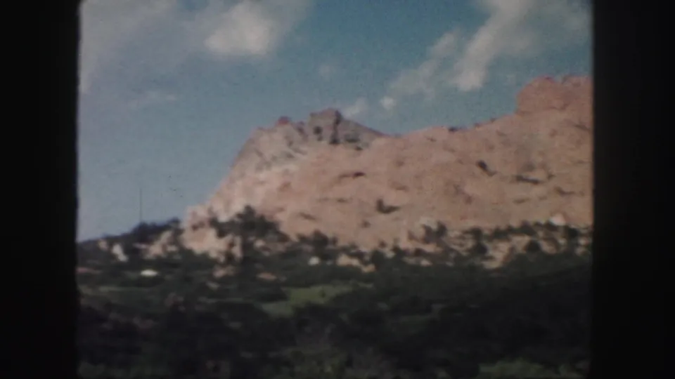 1958 Colorado travel footage frame with rocky tan ridge, green vegetation, and soft 8mm color rendering.