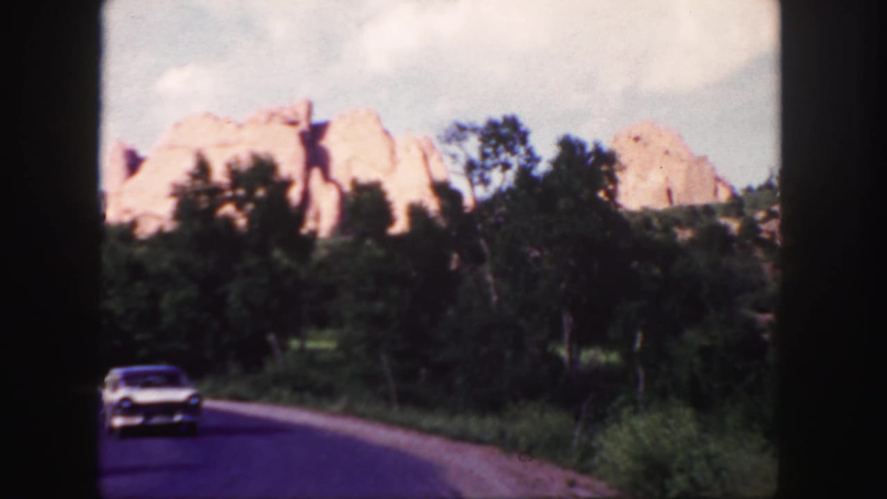1958 Colorado 8mm frame showing a white sedan approaching on a road with red rock formations and green trees behind it.