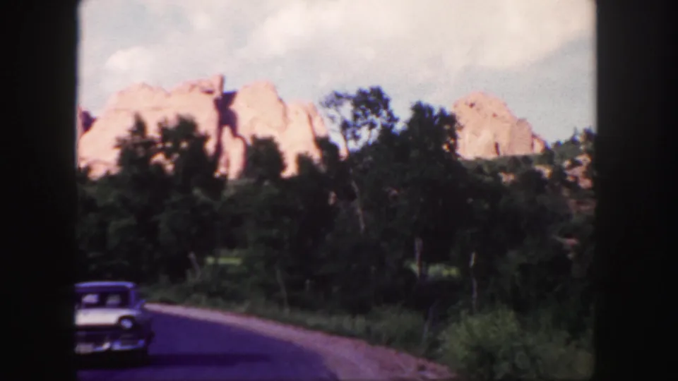 1958 Colorado home-movie frame of a white sedan moving past roadside trees with red rock formations beyond.