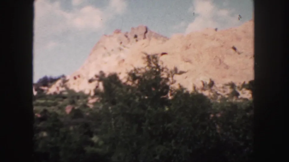 1958 Colorado 8mm frame showing a red rock hillside above green trees, with no vehicle visible in this frame.