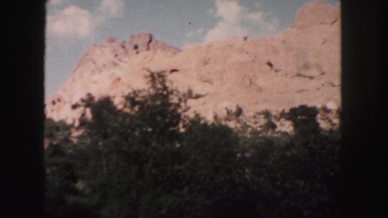 1958 Colorado 8mm frame showing reddish rock formations rising behind dark foreground trees and scattered clouds.