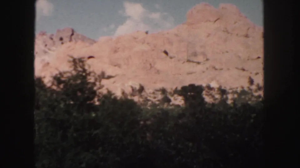 Colorado home-movie frame with sunlit reddish rocks, dark tree line, pale sky, and soft 8mm color grain.