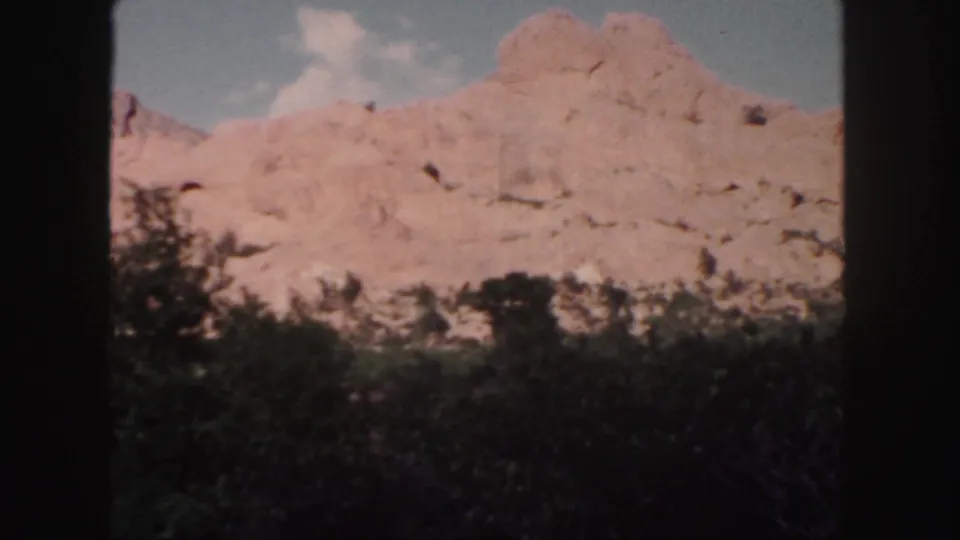 1958 Colorado landscape frame with pale red rock faces, foreground vegetation, and a cloudy blue sky in soft focus.