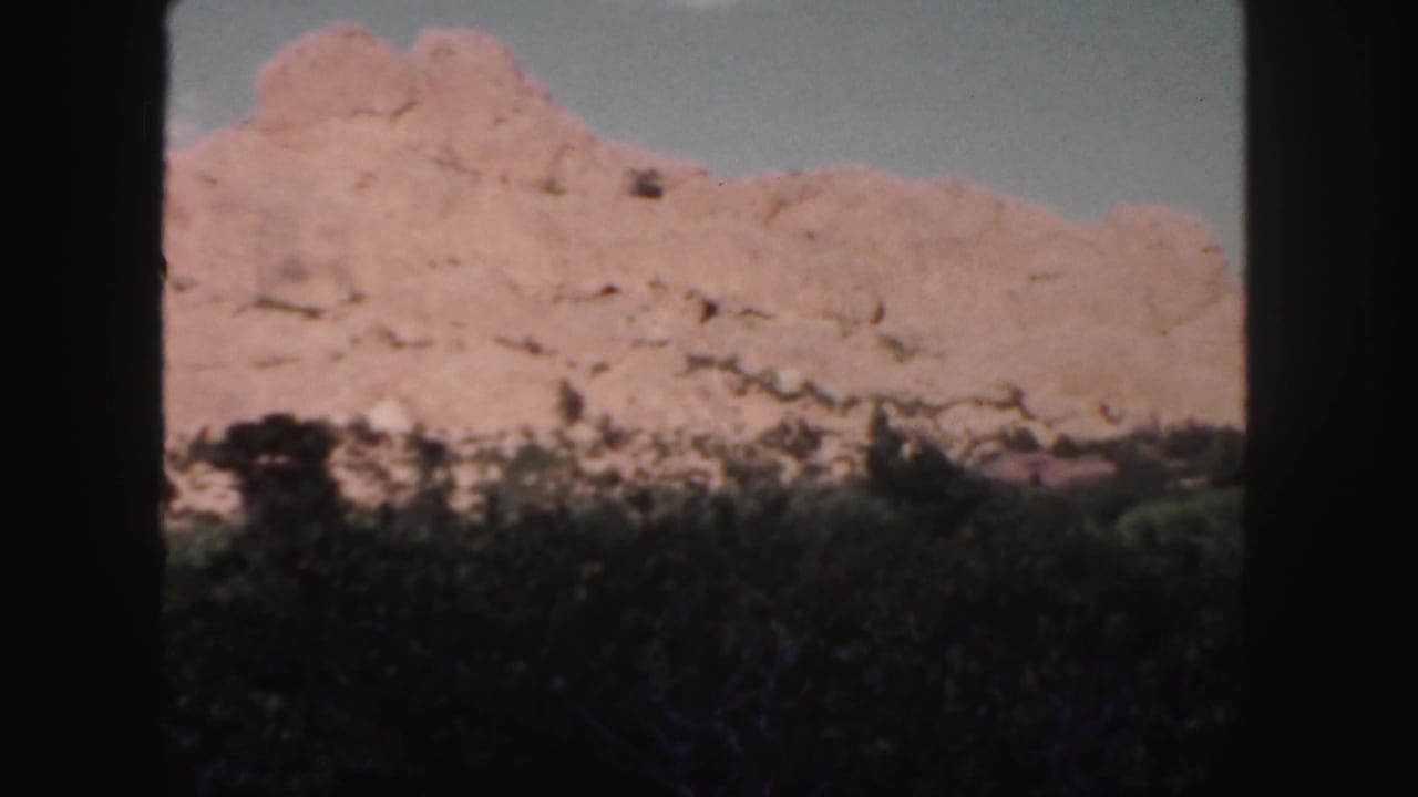 1958 Colorado home-movie frame showing a red rock ridge above dark shrubs and trees beneath a pale blue sky.
