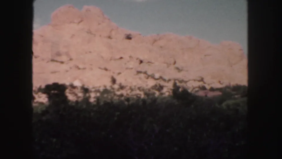 1958 Colorado home-movie frame showing a red rock ridge above dark shrubs and trees beneath a pale blue sky.