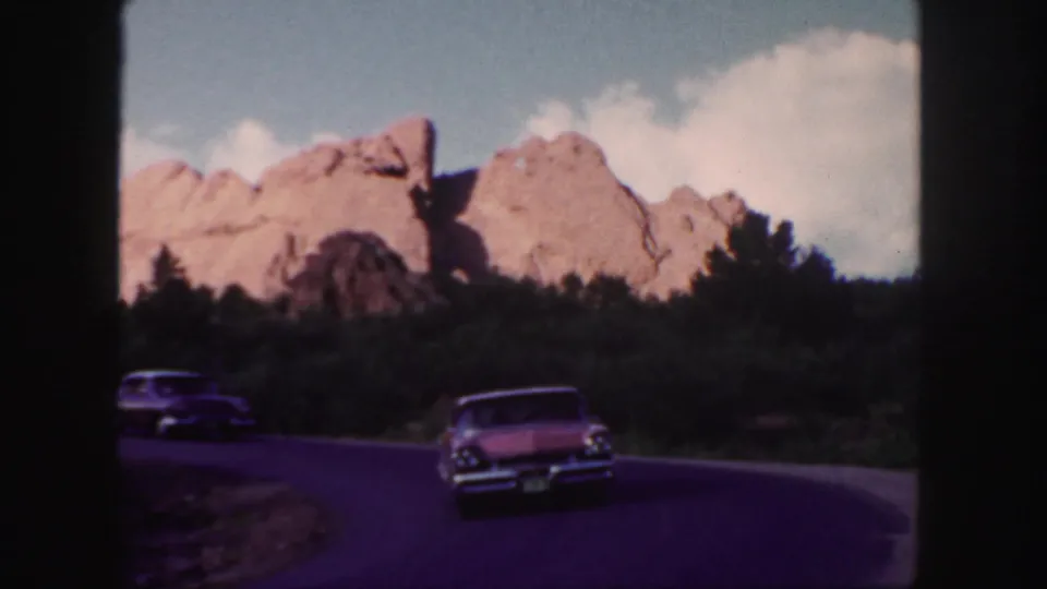 Classic cars travel along a curving Colorado road beneath red rock formations and a bright clouded sky.