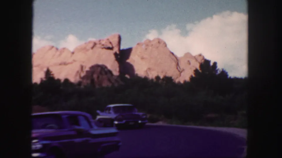 A classic car passes close to camera as another car approaches on a winding Colorado road below red rock scenery.