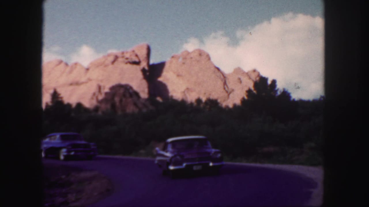 Two classic cars approach a curve below red rock formations and trees.