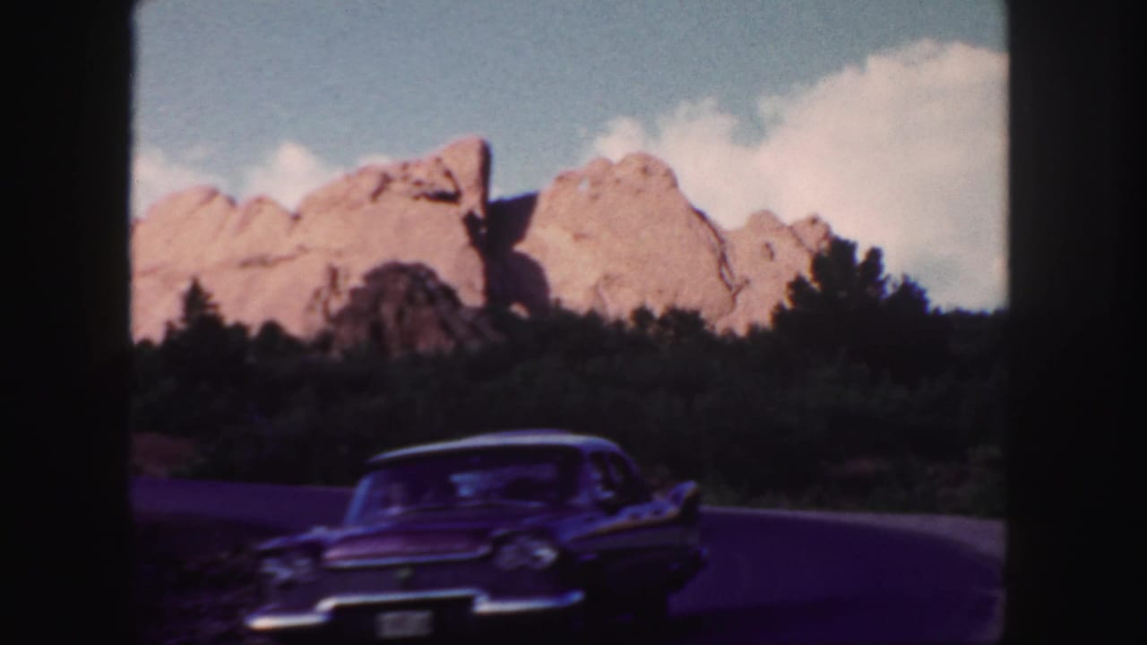 A dark classic car passes close to camera on a curved road near red rocks.