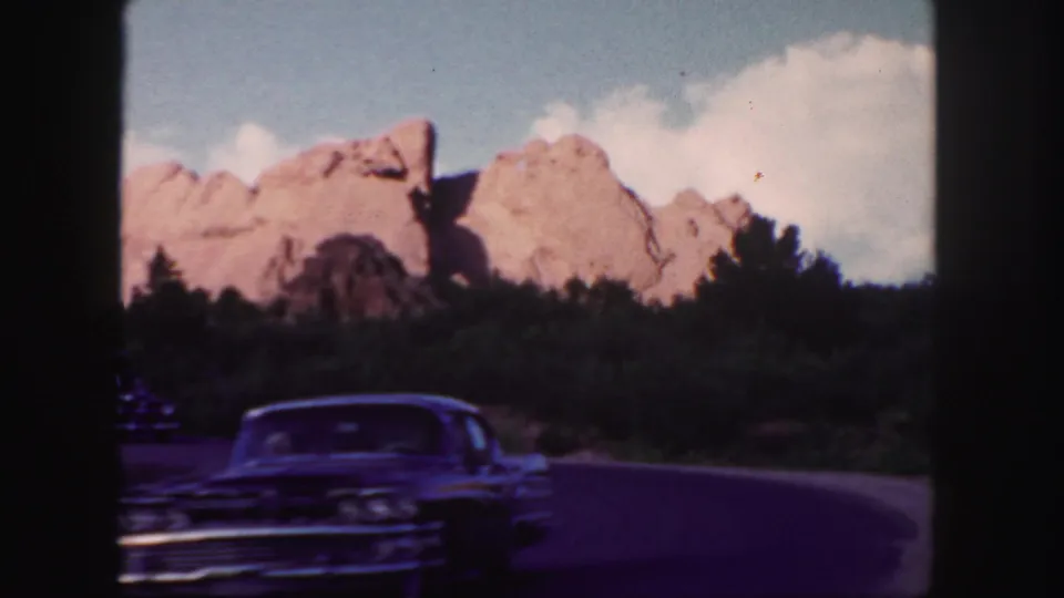 Soft 8mm frame from 1958 Colorado showing a dark classic automobile passing across a winding road with red rocks and trees behind it.