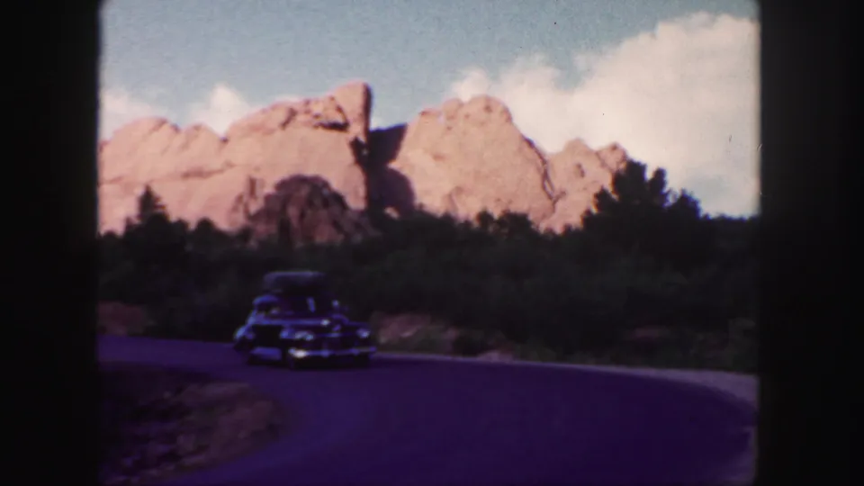 1958 Colorado 8mm frame showing a dark classic car farther down the bend, with roadside greenery and red rock formations behind it.