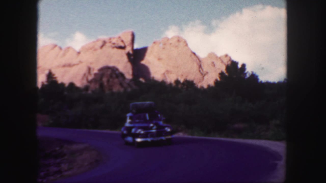 Vintage car on a curving road below pale red rock formations.