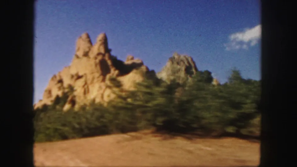 1958 Colorado travel footage frame with tan rock spires rising beyond trees and dry roadside terrain under a clear sky.