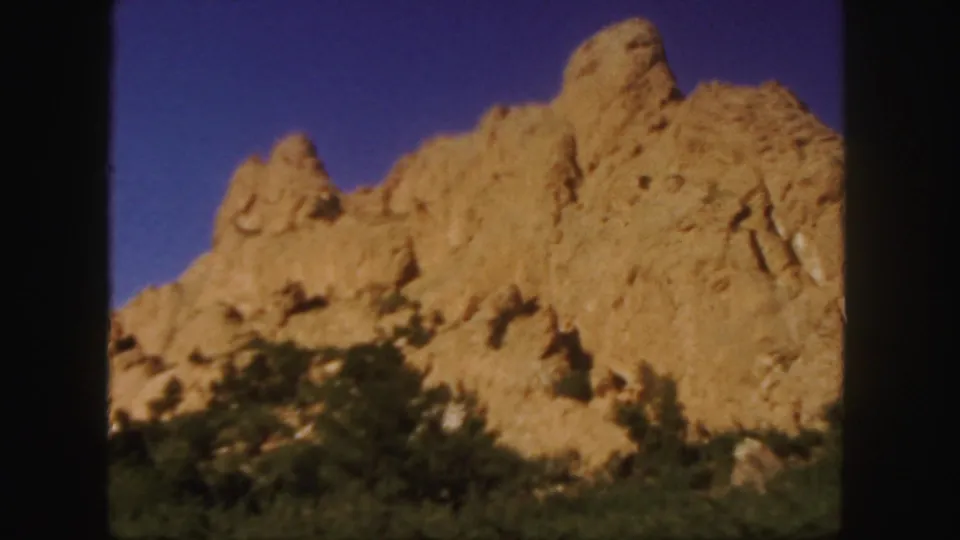 1958 Colorado 8mm frame showing a closer roadside look at a tan rocky ridge with shrubs at its base.