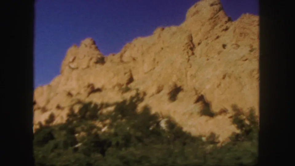 1958 Colorado home-movie frame with a tan rock wall and scrub vegetation blurred by motion from the moving vehicle.