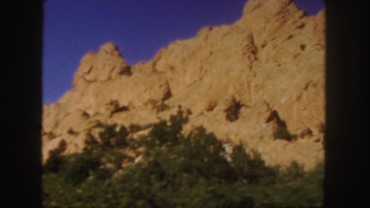 Tan rocky mountain ridge above dark green roadside vegetation under a deep blue sky.