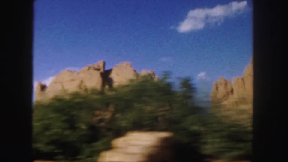 1958 Colorado 8mm roadside view with distant tan rock formations, blurred greenery, blue sky, and a small cloud.