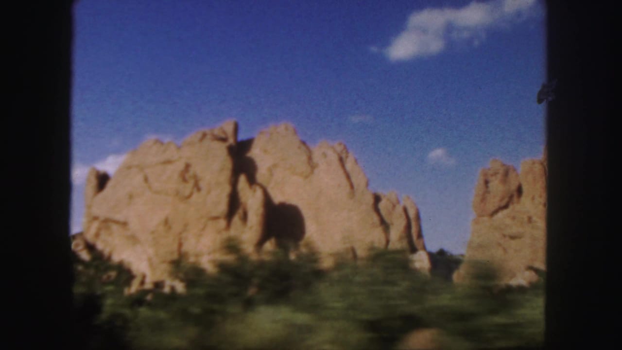 1958 Colorado 8mm frame showing a sandstone rock formation from a moving vehicle, with shrubs blurred in the foreground and blue sky above.