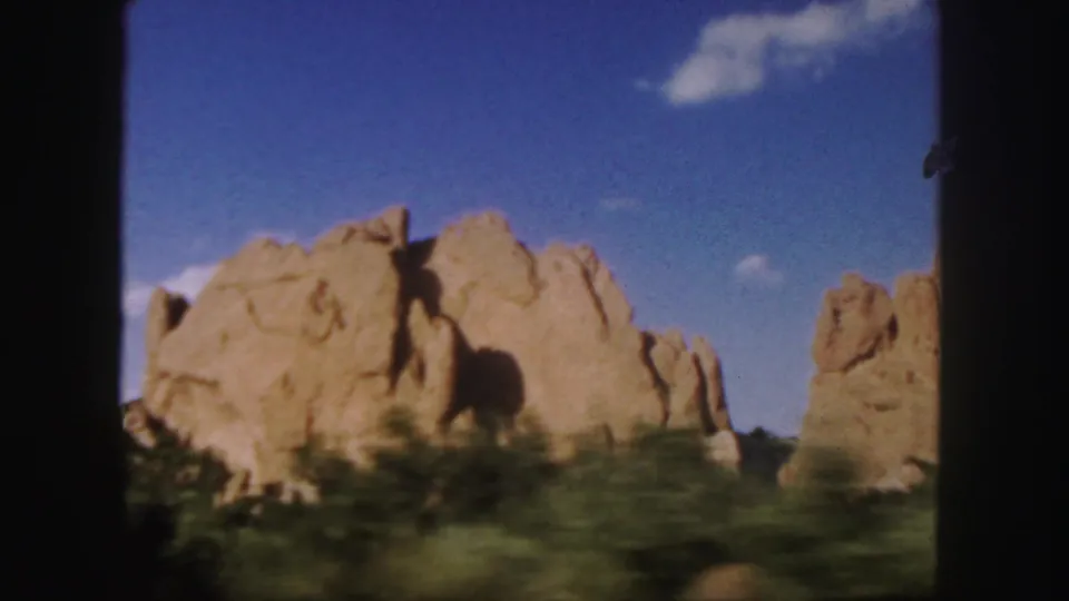 1958 Colorado 8mm frame showing a sandstone rock formation from a moving vehicle, with shrubs blurred in the foreground and blue sky above.