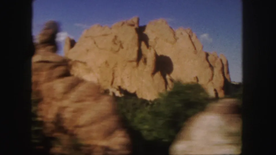 1958 Colorado 8mm frame with a blurred foreground rock column passing in front of tan sandstone formations and low green vegetation.