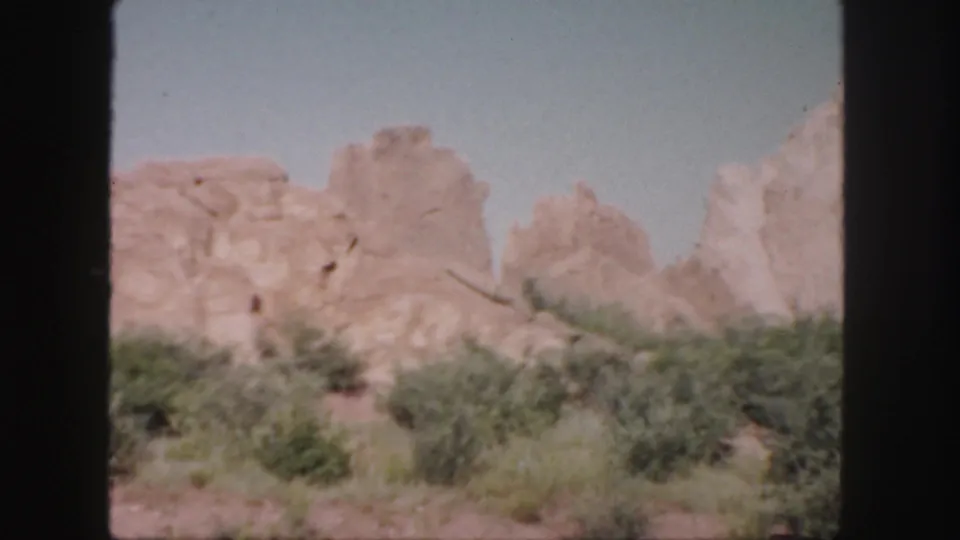 1958 Colorado 8mm frame with a brighter, paler exposure showing sandstone rock forms behind shrubs and grasses under a light blue sky.