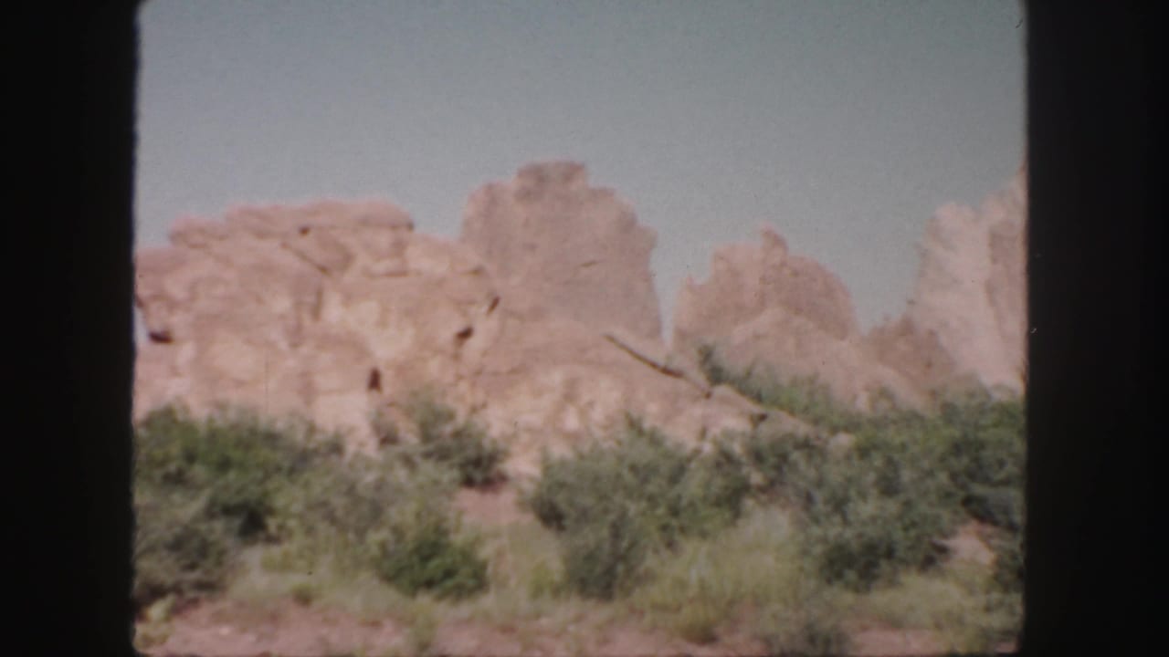 1958 Colorado 8mm frame showing pale sandstone outcrops rising beyond low scrub and dry ground.