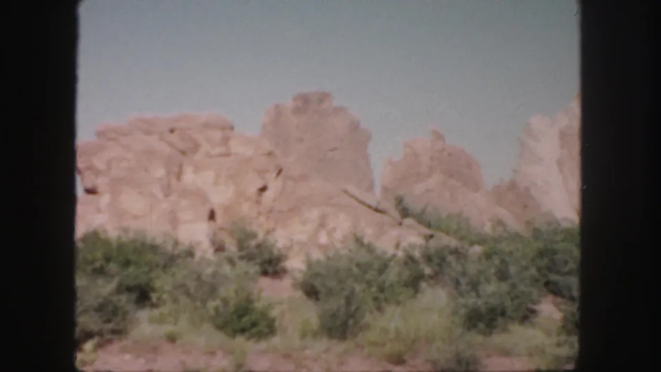 1958 Colorado 8mm frame showing pale sandstone outcrops rising beyond low scrub and dry ground.