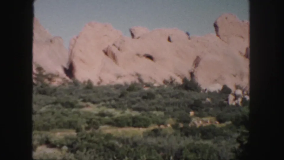 1958 Colorado 8mm frame showing pale pink sandstone formations rising behind dense green desert vegetation under clear sky.