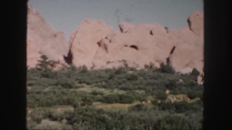 1958 Colorado landscape frame with soft-focus pink sandstone outcrops, green brush, and clear sky in vintage 8mm color.