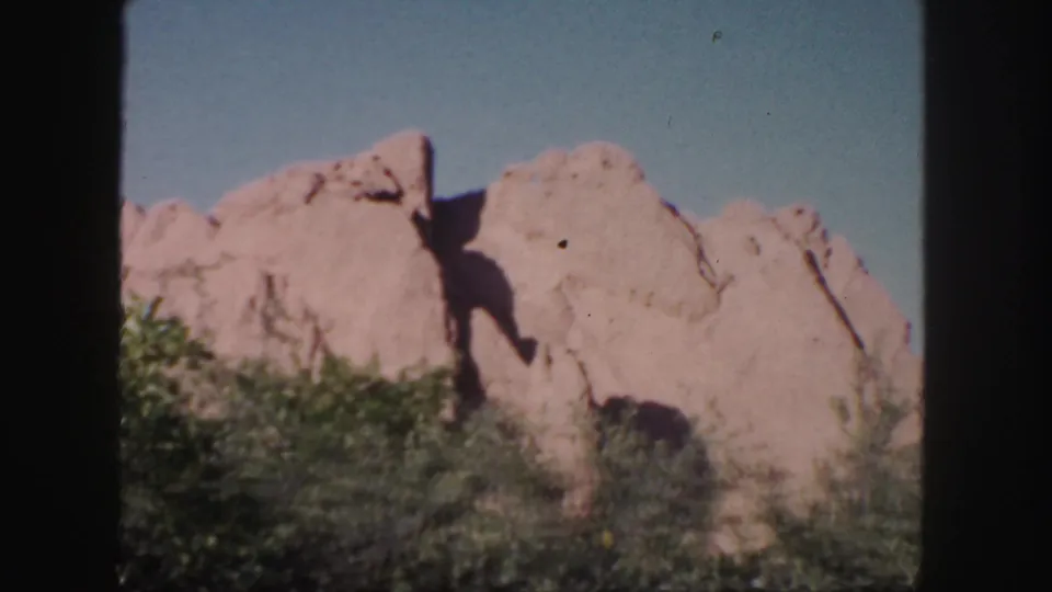 Colorado 8mm frame showing a large pink sandstone formation, deep rock shadow, foreground shrubs, and clear blue sky.