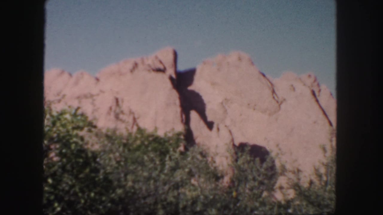 1958 Colorado 8mm frame showing a sunlit tan rock face rising behind thick green shrubs under blue sky.