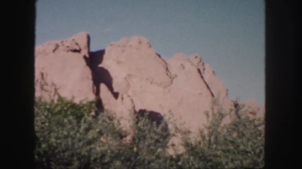 1958 Colorado home-movie frame showing a sunlit rocky slope above shrubs, with dark 8mm frame borders visible.