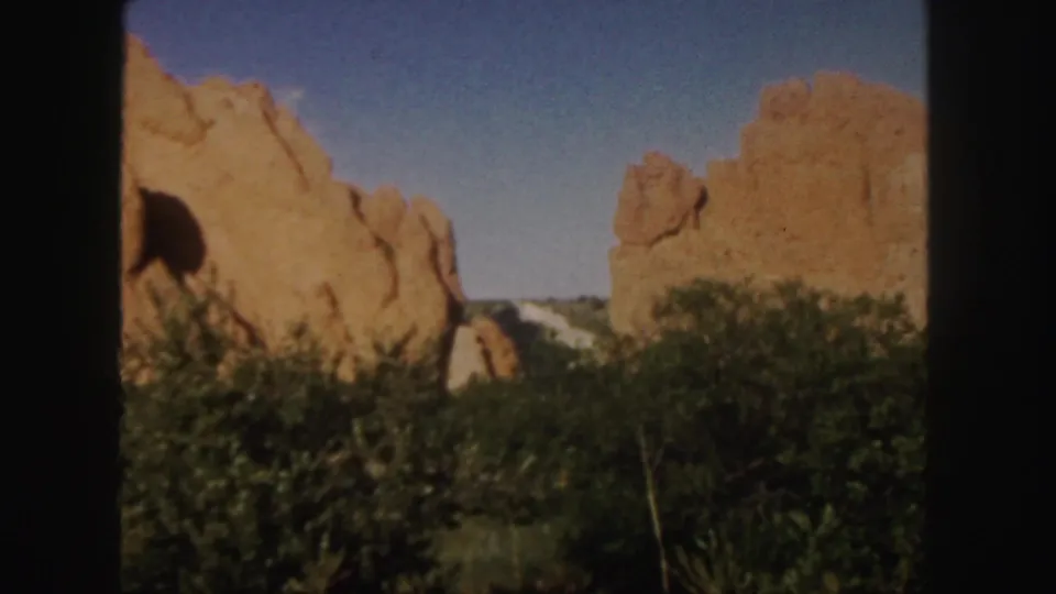 1958 Colorado 8mm frame showing warm red rock formations, dense shrubs, and a clear blue sky from a ground-level viewpoint.