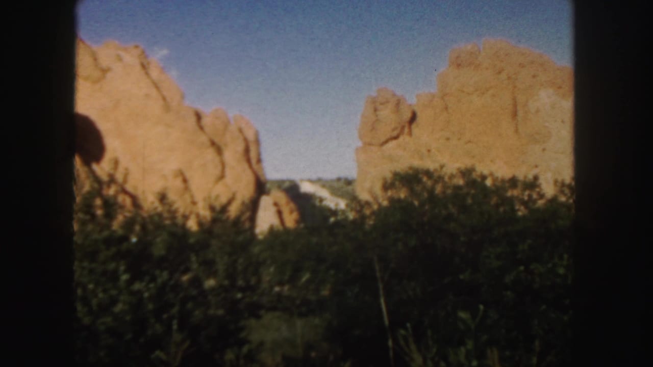 1958 Colorado 8mm frame showing tan-red sandstone formations, green shrubs, blue sky, and dark film borders.