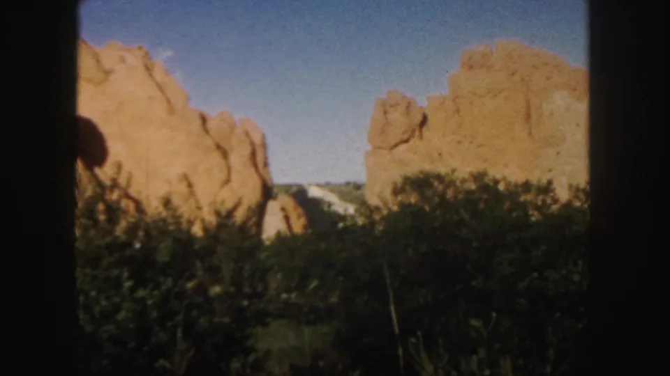 1958 Colorado 8mm frame showing tan-red sandstone formations, green shrubs, blue sky, and dark film borders.