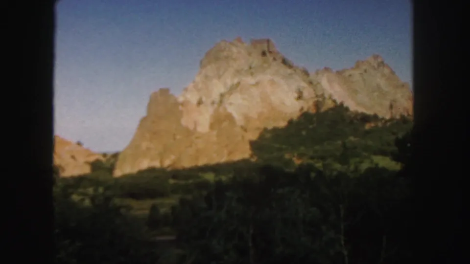 1958 Colorado 8mm frame with a pale red rock formation, blue sky, and dense foreground vegetation.