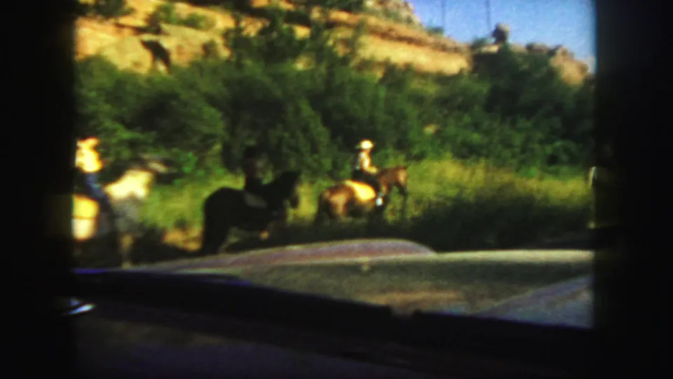 Two distant riders in hats move on horseback through green Colorado vegetation, with a vehicle edge visible in the foreground.